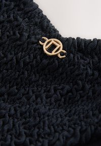Tissu noir texturé avec un petit emblème métallique doré rond présentant un motif géométrique fixé près du bord supérieur.