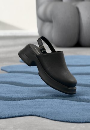 Sabot en cuir noir avec sangle réglable présenté au-dessus d'un tapis bleu texturé sur un sol gris dans un environnement intérieur minimaliste.
