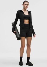 Kvinde i sort sports-bh med prikker, jakke og shorts, som holder en rullet yogamåtte, iført sorte strømper og slip-on sko, stående på en hvid baggrund.