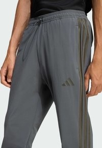 Šedé športové nohavice s elastickým pásom a šnúrkou, s tromi bočnými pruhmi v olivovej farbe, hladkým povrchom a logom Adidas v tmavozelenej farbe.