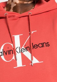 Gros plan d'un sweat à capuche corail avec le logo "Calvin Klein Jeans" et des cordons, porté par une personne avec du vitiligo sur le cou.