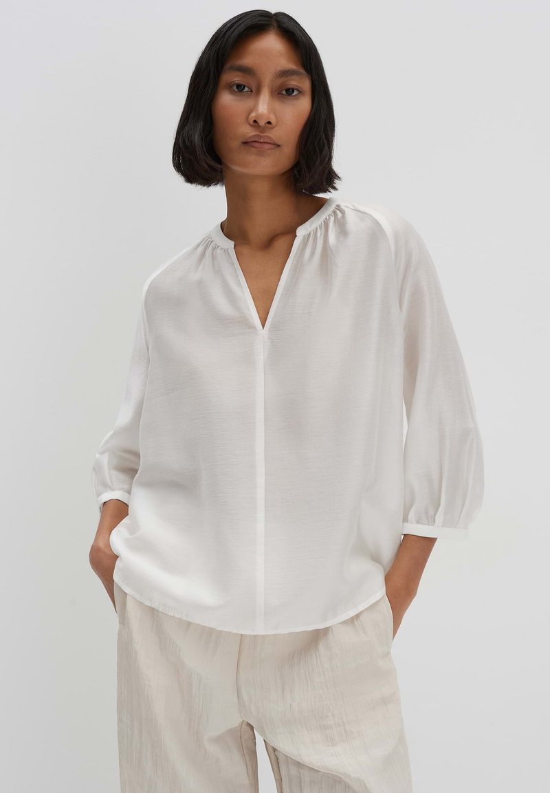 Blusa bianca con scollatura a V, maniche lunghe a sbuffo e leggere arricciature sul collo, realizzata in tessuto leggero e testurizzato.