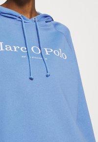 Felpa di colore azzurro chiaro con cappuccio, dotata di un cordoncino, polsini a coste e il nome del marchio "Marc O Polo" in bianco sul davanti. Tessuto morbido e texturizzato.