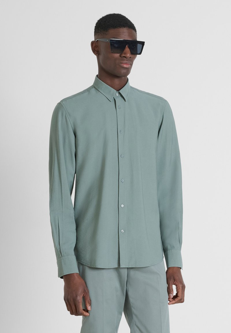 Camicia a maniche lunghe con bottoni in morbido tessuto verde tenue, caratterizzata da un colletto classico e chiusura con bottoni, abbinata a grandi occhiali da sole neri.