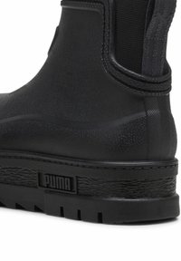 Gros plan sur une bottine Puma noire avec une semelle en caoutchouc texturée, un panneau latéral élastique et un logo de marque sur le talon.