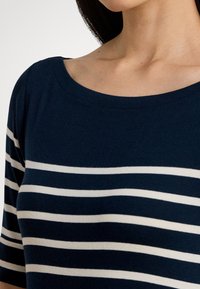 Γυναίκα που φοράει μπλε ναυτικό πουκάμισο με λευκές οριζόντιες ρίγες και φαρδιά στρογγυλή λαιμόκοψη, κομμένο στους ώμους και το πάνω μέρος του στήθους.