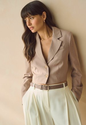 Blazer corto marrone con bottoni dorati, abbinato a pantaloni plissettati color crema. Completato con una cintura beige e tocchi di gioielleria dorata minimal.