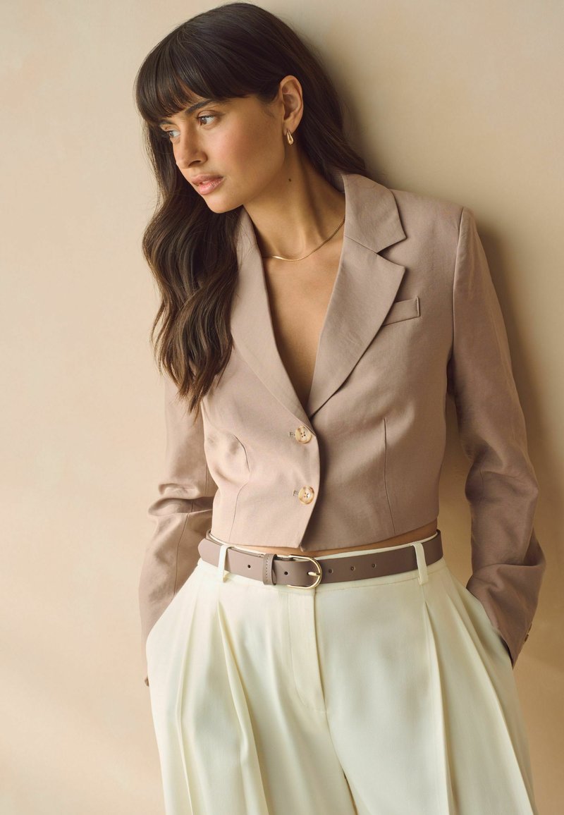 Blazer court marron avec des boutons dorés, assorti à un pantalon plissé crème. Complété par une ceinture beige et des accents de bijoux en or minimalistes.