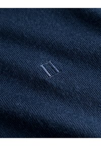 Marineblauer Strickstoff mit sichtbarer Textur, versehen mit einem kleinen gestickten "II" in einem etwas helleren Farbton. Weiches, gleichmäßig gewebtes Material.