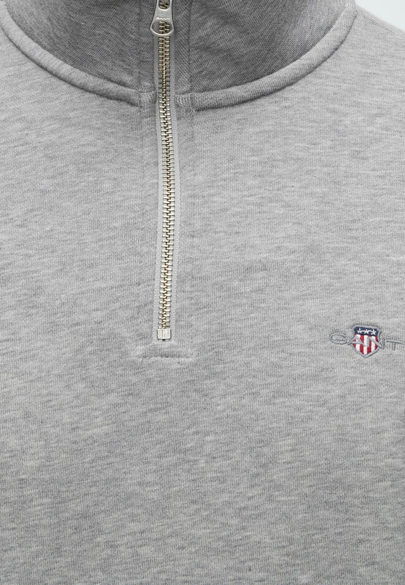Hellgrauer Viertel-Zip-Sweatshirt mit silbernem Reißverschluss, hohem Kragen und kleinem gesticktem Schild-Logo in Rot, Weiß und Blau auf der Brust.