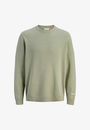 Dječak s bež kapom, slojevitim svijetlozelenim džemperom, bijelom košuljom, tamnoplavom traper jaknom i hlačama te bež cipelama stoji na bijeloj pozadini.