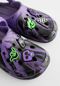 Violet un melnas koka kurpes ar vairākiem dekoratīviem gumijas piekariņiem, tostarp skeletu, liesmu un šaha karogu, ar ventilācijas caurumiem visā virsmā.