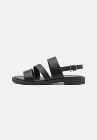 Sandalias de cuero negro con tres correas anchas, correa ajustable en el tobillo, suela plana y diseño minimalista sobre un fondo blanco.