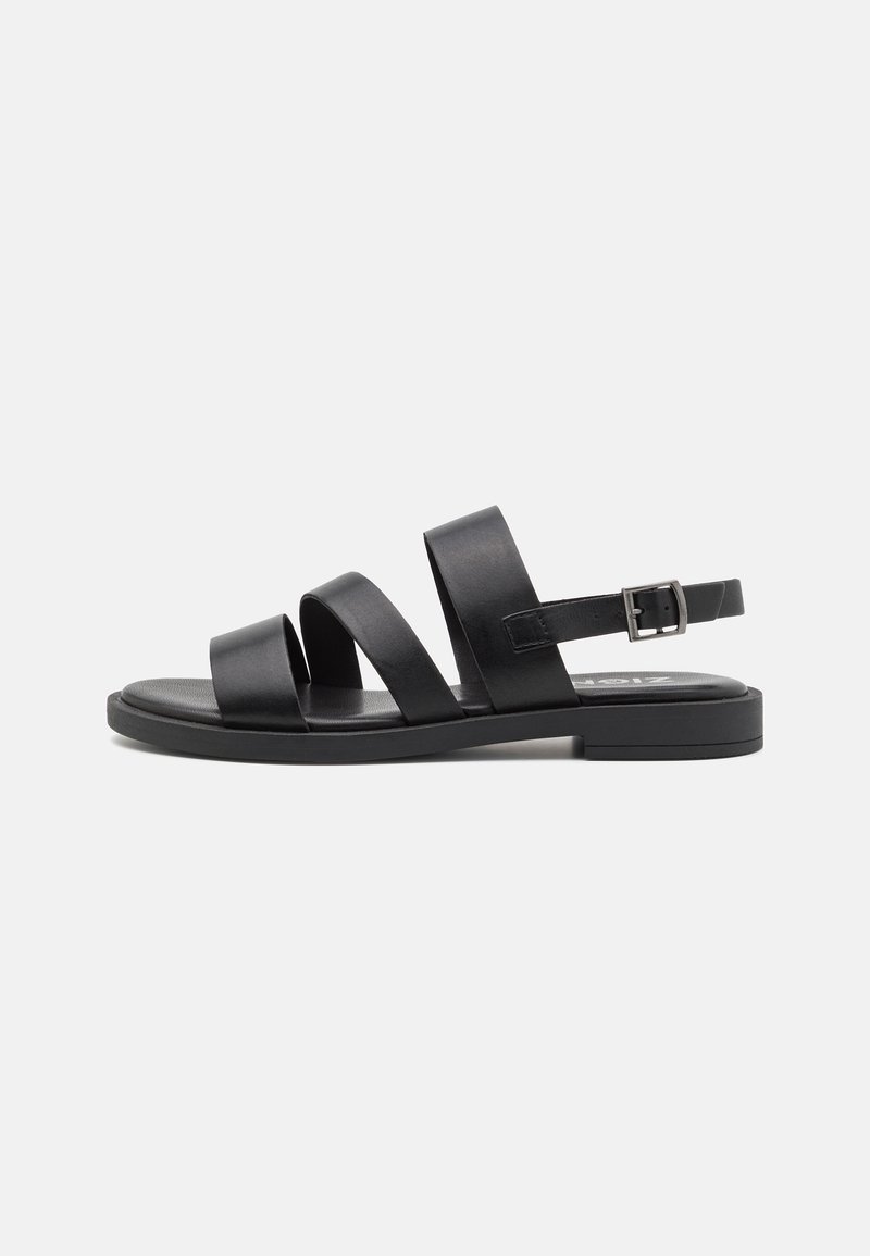 Sandalias de cuero negro con tres correas anchas, correa ajustable en el tobillo, suela plana y diseño minimalista sobre un fondo blanco.