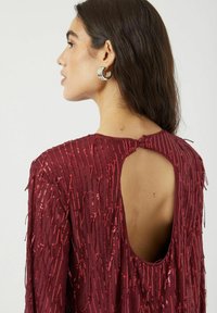 Viininpunainen mekko, jossa on teksturoitu pinta, hapsutetut paljetit ja avonainen selkäosa. Pitkät hihat ja niskassa nappikiinnitys.