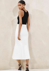 Sort ribbet tanktop parret med en hvid, mellemlang, flydende nederdel. Modellen har sorte hæle sandaler på. Glat, minimalistisk design.