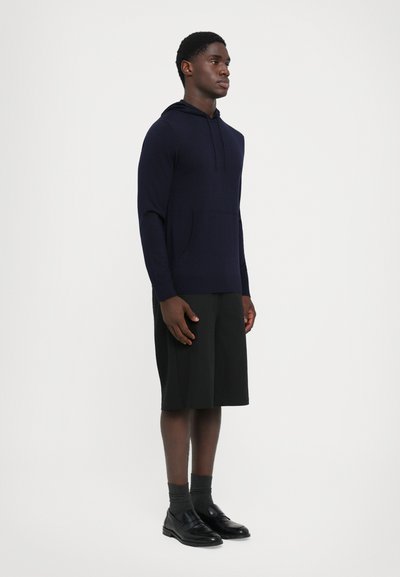 Felpa a maglia blu navy con tasca frontale, abbinata a pantaloni corti neri fino al ginocchio, calzini grigi e scarpe nere senza lacci, in piedi su uno sfondo uniforme.