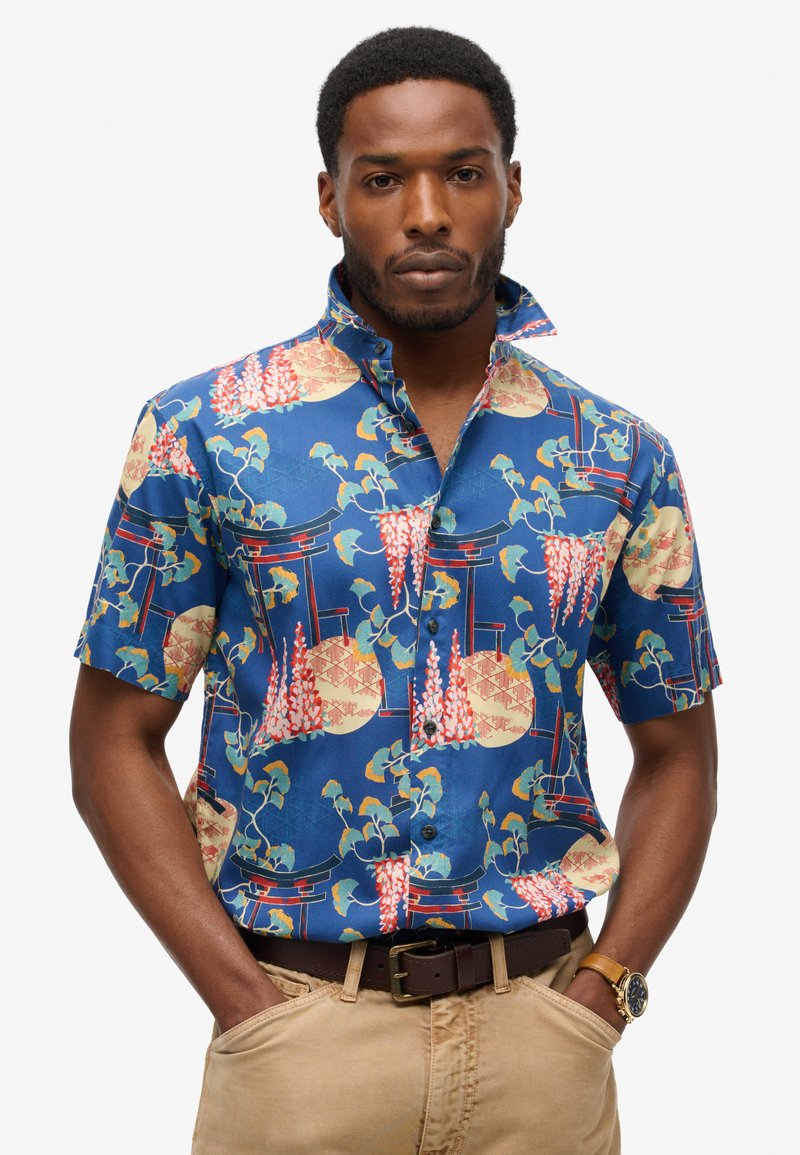 Korte mouwen blouse in blauwe stof met kleurrijke bloemen- en architectonische patronen, voorzien van een kraag en een borstzakdetail.