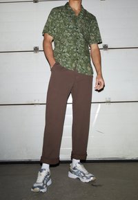 Man som bär en kortärmad skjorta med grön lövmönster, bruna upprullade byxor, vita strumpor och vit-grå sneakers, stående inomhus.