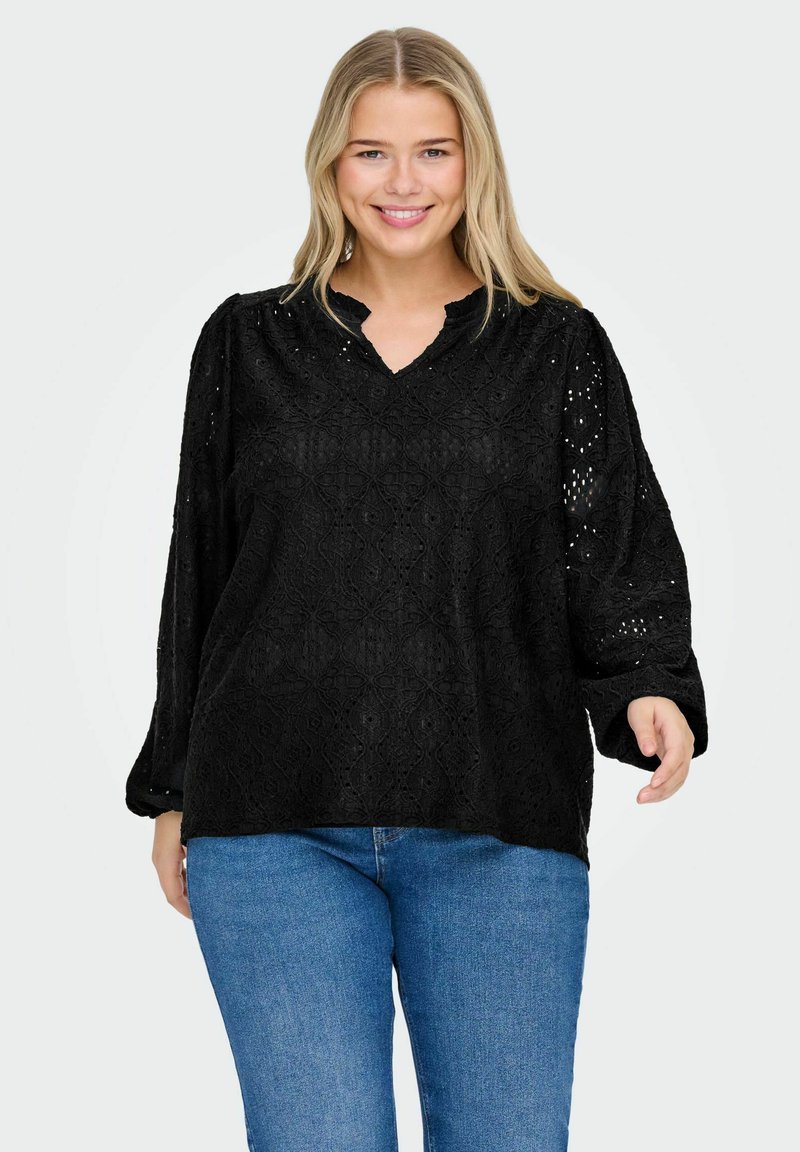 Blusa in pizzo nero con scollo a V, caratterizzata da motivi floreali tridimensionali, maniche lunghe a palloncino e accenti di tessitura aperta. Indossata con jeans blu.