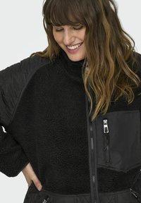 Black fleece jacket with a high collar, quilted fabric accents, zippered pockets, and a soft, textured surface. Durable and warm design.