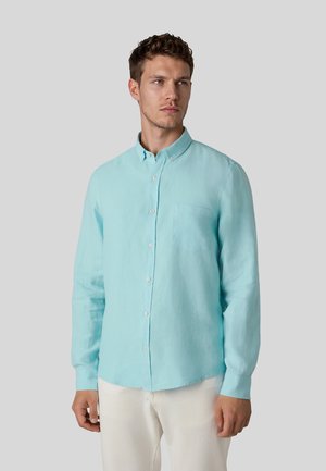 Mann trägt ein hellblaues, langärmeliges Hemd mit Knöpfen und weiße Hose, schaut leicht zur Seite vor einem einfarbigen grauen Hintergrund.