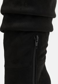 Bota de ante negra hasta la rodilla con la parte superior doblada, cierre de cremallera a lo largo del lado y una textura suave.