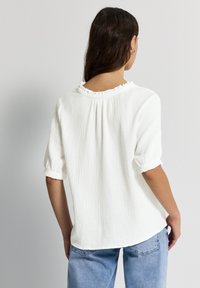 Haut blanc texturé avec un col à volants, des manches courtes retroussées et une coupe ample. Porté avec un jean bleu clair, vu de dos.
