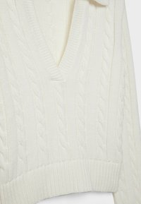 Gilet en maille torsadée blanche avec col en V et ourlet côtelé, montrant des coutures détaillées et un tissu doux texturé.