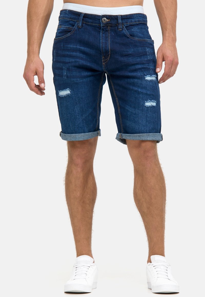 Homme portant un short en jean bleu foncé roulé avec des patches usés et des baskets blanches, ceinture blanche visible.