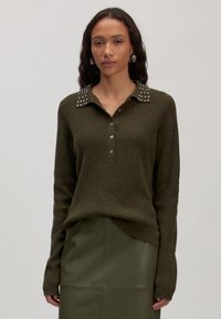 Vrouw in een olijfgroen gebreid truitje met een studded kraag en knopen, gecombineerd met een bijpassende olijfgroene leren rok, tegen een effen achtergrond.