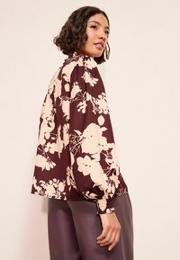 Bordová blúzka s kvetinovým vzorom v krémovej farbe, s vysokým golierom a balónovými rukávmi. Štruktúrovaná látka s zhromaždenými manžetami a voľným strihom.