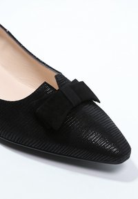 Chaussure plate noire à bout pointu, matériau texturé, avec un nœud en daim sur le dessus. Doublure intérieure lisse.