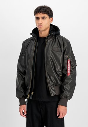 Junger Mann trägt eine dunkelbraune Leder-Bomberjacke mit Fellkragen, einer aufgenähten Brustemblem und einem roten "Remove Before Flight"-Anhänger an der Ärmeltasche.