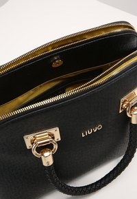 Zwarte textuur leren handtas met gouden hardware, ritssluiting en gevlochten handvat. Het interieur heeft een contrasterende gouden voering. Merkennaam "LIU JO" zichtbaar.