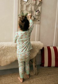 Pigiama verde menta con un motivo di alberi di Natale rosa e dorati. Il bambino sta in piedi su un tappeto texturizzato, allungandosi per prendere un oggetto appeso.