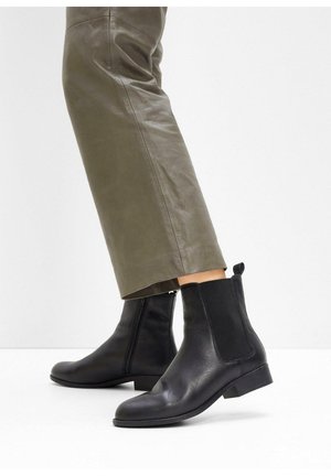 Stivali Chelsea neri in pelle con pannelli laterali elastici e un pratico passante. Texture liscia e punta arrotondata, abbinati a pantaloni in pelle verde oliva.