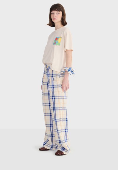 Femme portant un t-shirt graphique beige, un pantalon large à carreaux, un chouchou à carreaux bleus au poignet et des sandales marron, debout devant un fond blanc.