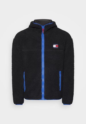 Veste en polaire noire avec une surface texturée, zip bleu et accents, capuche et un petit patch logo sur la poitrine. Deux poches latérales.