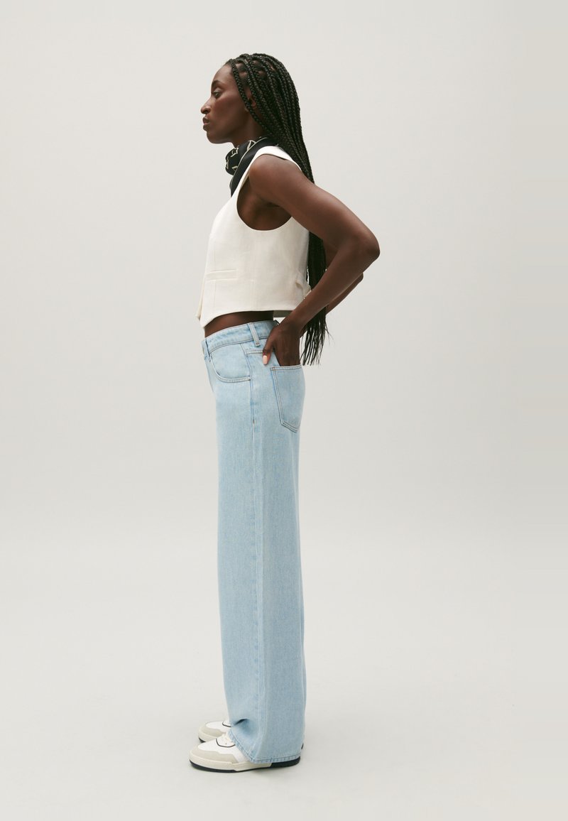 Gilet en lin blanc raccourci associé à un jean en denim large de couleur bleu clair. Tenue complétée par des baskets blanches et grises et un accent avec une écharpe noire.