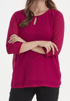 Blusa fucsia con maniche a tre quarti, scollatura rotonda e dettaglio a chiave. Tessuto leggero e trasparente con una texture liscia. Pantaloni neri.