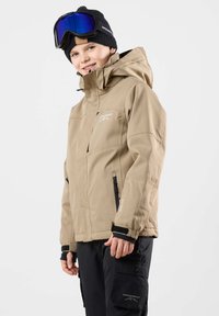 Beige wasserdichte Jacke mit Reißverschlusstaschen, verstellbaren Bündchen und Kapuze, kombiniert mit schwarzen Cargo-Hosen. Schwarze Mütze und blaue Skibrille.