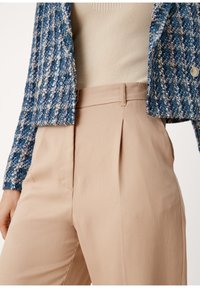 Blå och gräddfärgad tweedjacka med en strukturerad mönstring, kombinerad med beige veckade byxor. Fokusera på tyg och färgkontrast.