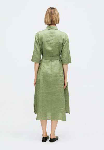 Γυναίκα με κοντά ξανθά μαλλιά, που φοράει ένα ελαιί πράσινο φόρεμα μέχρι το γόνατο με ζώνη και μανίκια μέχρι τους αγκώνες, εμφανιζόμενη από πίσω, στέκεται σε λευκό φόντο.