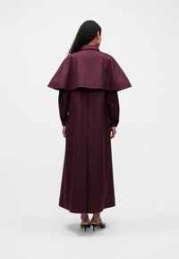 Sieviete ar cirtainiem matiem, ģērbta garā purpura mētelī ar apmetni aizmugurē un melnām augstpapēžu kurpēm, stāv uz baltā fona.