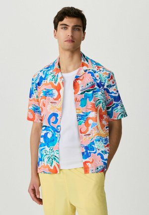 Młody mężczyzna ubrany w kolorową koszulę z krótkim rękawem w tropikalny wzór, na białym T-shircie i żółte krótkie spodenki, stoi na prostym tle.