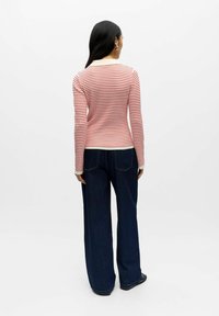 Moteris, stovinti atsukusi nugarą, dėvi kreminės ir raudonos spalvos dryžuotą ilgomis rankovėmis megztinį, tamsius plačius džinsus, juodus batus ir sidabrines apvalias auskarus.