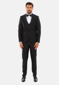 Tuxedo nero con rever in satin, camicia bianca e cravatta a farfalla nera. Scarpe nere lucide. Design slim fit, in piedi su uno sfondo bianco.
