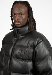 Black puffer jacket with a high collar, made of smooth leather. Features a textured logo on the chest and a zip closure.