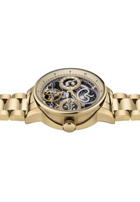 Montre en or avec un cadran squelettique, montrant les mécanismes internes. Dotée d'un bracelet en métal lisse et d'une couronne centrale pour les réglages.